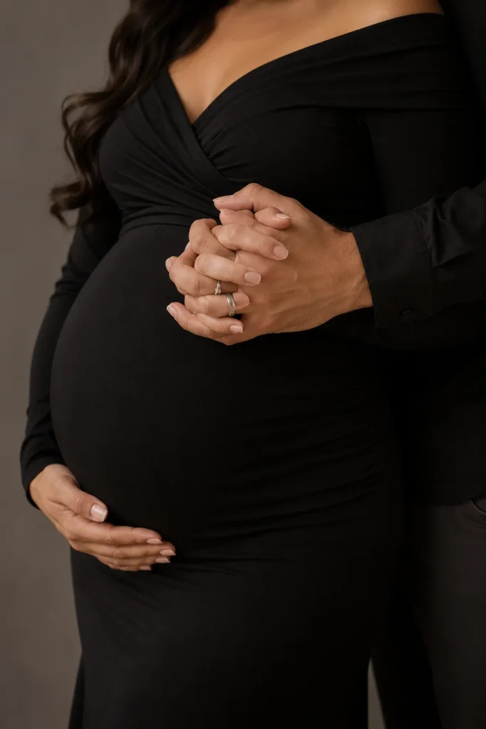 Couple gently interlocking their fingers above a pregnant belly in an elegant studio maternity portrait.